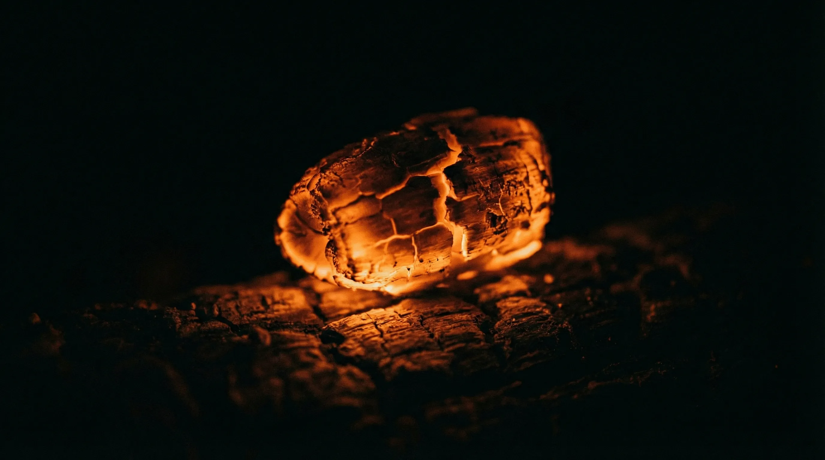 A single glowing ember on charred wood, its cracked surface radiating warm amber light against pure darkness
